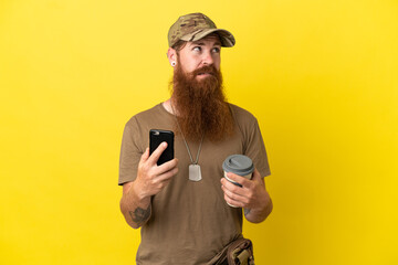 Redhead Military man with dog tag isolated on yellow background holding coffee to take away and a mobile while thinking something