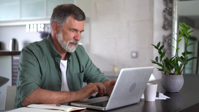 Bearded man working online with laptop computer at home sitting at desk. Home office, browsing internet.
