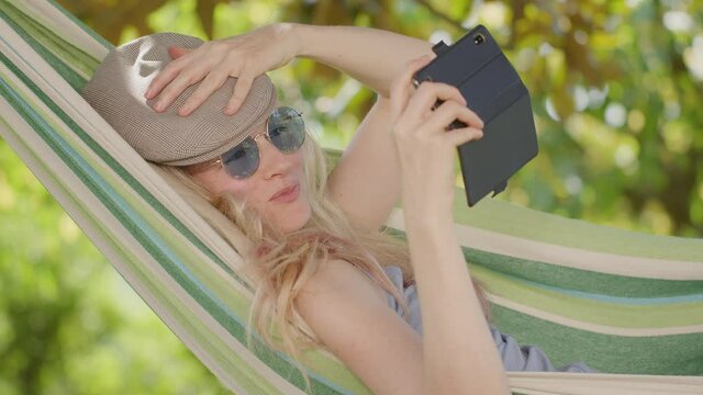 Smiling blonde woman with sunglasses using smartphone, lying relaxing on the hammock in the garden, free time and summer holiday concept to surf the internet or chat with friends using social media