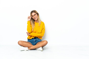 Young blonde Uruguayan woman sitting on the floor isolated on white background with glasses and happy