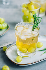 Refreshing summer drink in glass with ice cubes, green grape and rosemary on the table.