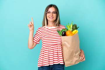 Young woman holding a grocery shopping bag isolated on blue background pointing up a great idea