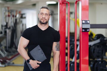 Muscular handsome trainer looking holding a fitness plan on clipboard for working out in the fitness gym while smiling at camera