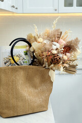 Stylish beach bag with beautiful bouquet of dried flowers and magazine on white table in kitchen