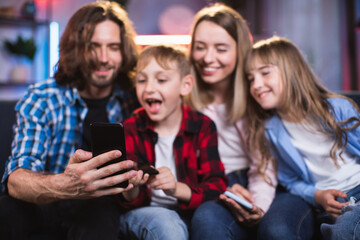 Playful kids and their young parents sitting in hugs on couch and taking selfie on modern cell phone. Concept of family, technology and leisure time.