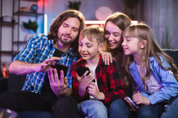 Smiling parents with son and daughter using mobile application for entertaining during evening time. Caucasian family with gadgets at home.