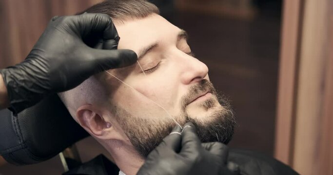 Barber doing threading procedure on face of man at salon
