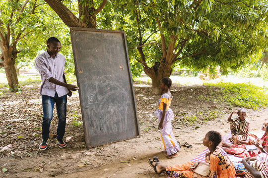 Tall black teacher in casual clothing inviting one of his small pupils to write something on the blackboard in an open air school in West Africa