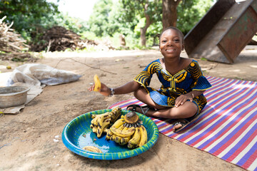 Gentle little black African girl sitting on a coloured mat in the middle of a rural courtyard,...