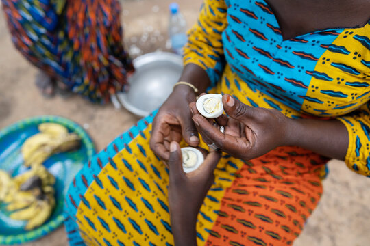 Caring African woman in a colorful dress sitting on the floor in a rural village kitchen, sharing half of an egg with a child; poverty concept