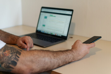 Close-up of a young Latino man's arm and hand using a cell phone with a laptop in the background