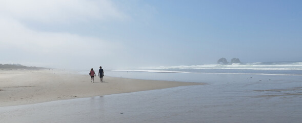 Misty Beach Walk Panorama- A couple walks the Rockaway Oregon beach in the morning before the mist has burn off. Twin Rocks sea stacks sit on the horizon.