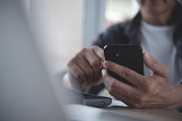 Asian man using mobile smart phone during online working on laptop computer on table in modern home office