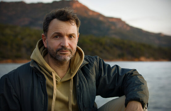 Handsome middle aged man sitting by the sea in the evening with mountains in the background. The Hazards range Freycinet National Park Tasmania Australia