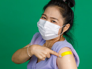 Studio close up shot of Asian female patient with face mask look at camera pointing at yellow bandage plaster after receive coronavirus covid 19 vaccination from doctor at clinic on green background