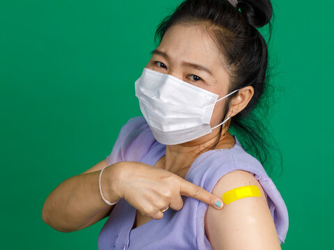 Studio Close Up Shot Of Asian Female Patient With Face Mask Look At Camera Pointing At Yellow Bandage Plaster After Receive Coronavirus Covid 19 Vaccination From Doctor At Clinic On Green Background