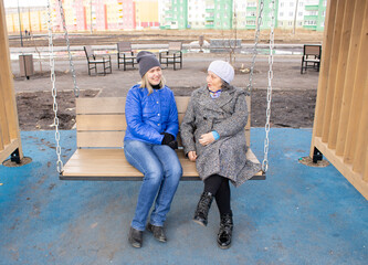 Two women sit on a swing in cold weather. Elderly woman and middle-aged woman outdoors.