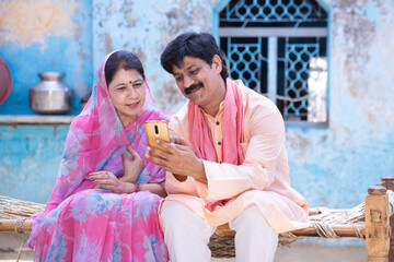 Happy rural indian couple using smart phone sit on bed. farmer husband wife watching at mobile screen, man woman smiling.Internet video call,village people using cellphone Technology. digital india.