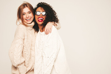 Two young beautiful smiling international hipster female wearing warm winter sweaters. Sexy carefree women posing on white background in studio. Hugging and going crazy. Happy and cheerful