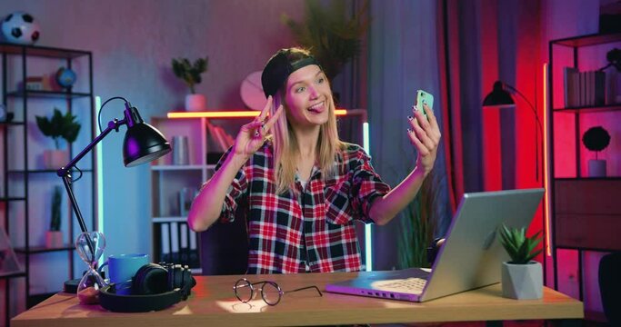 Adorable smiling happy young modern blondie with earring in nose having videocall on mobile with friend or relaitve when sitting at the desk at home in the evening
