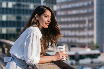 horizontal portrait of an attractive young indian business woman laying on a balcony railing. She holds a reusable coffee mug and has offices buildings on the background