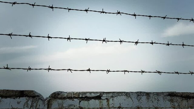 Looking at a wall with rows of barbed wire on top, with knots. Silhouetted handheld shot at sunset.
