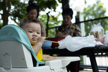 Asian boy sits in a chair with chopsticks in his mouth and is eating with his parents.