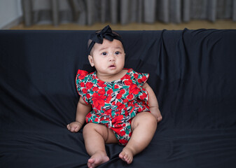 Fototapeta premium Little adorable baby girl sitting on black sofa alone. Asian 5 months old baby dressed in red dress with black headband sitting alone on sofa chair. 