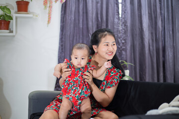 Happy mother and daughter sitting on sofa and playing. Mom and baby girl wearing same color dress. Lifestyle portrait of asian family. 