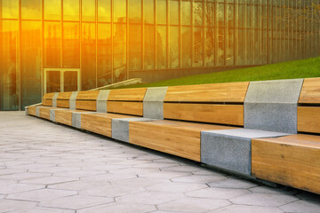 Wooden bench and tiled walkway. Office building for the work of company employees. Glass walls of a building with construction site reflection. Interior elements. Modern urban landscape.