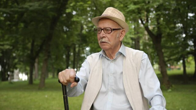 An old grandfather sits in the park on a bench and waits his grandson.