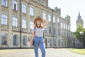 Cute happy female university or college student portrait. Studying abroad or educational concept. Cheerful young blonde with backpack and laptop wearing jeans and hat in campus. High quality image
