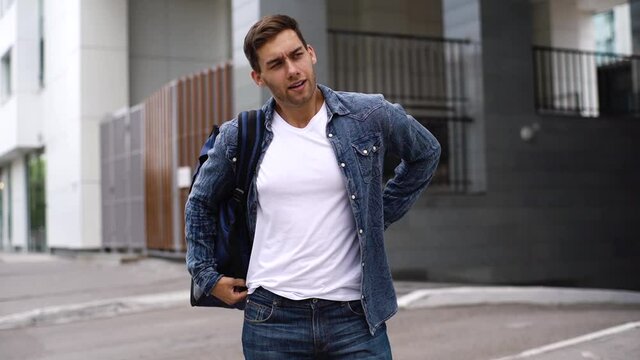 Handsome young delivery service employee putting large thermo backpack on shoulders walking down city street on summer day. Courier male ready delivery food to next client. Shooting in slow motion.