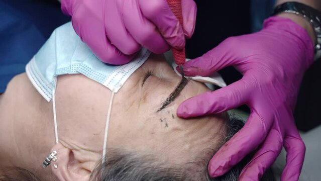 Cosmetology artist in sterile pink gloves drawing permanent eyebrows contour special make-up tattoo machine, beautician preparing middle-aged woman's face for microblading procedure in beauty salon