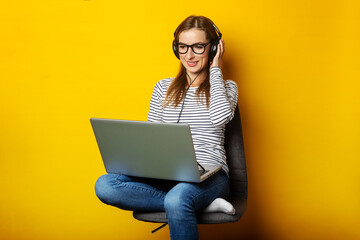 Young woman in headphones listening to music and sitting on chair and holding laptop on isolated yellow background