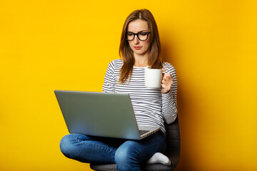 Young woman holding a cup of coffee, sitting on a chair, working on a laptop on an isolated yellow background