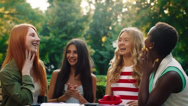 Four astonishingly beautiful women are sitting at a cafe table outside in a scenic park, conversing together and being joyful