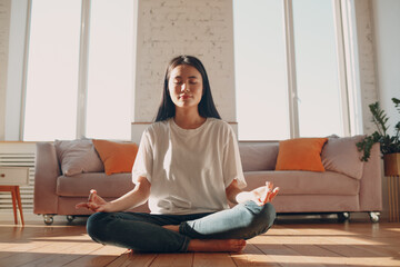 Asian woman doing yoga and zen like meditation lotus pose in casual wear at indoor living room...
