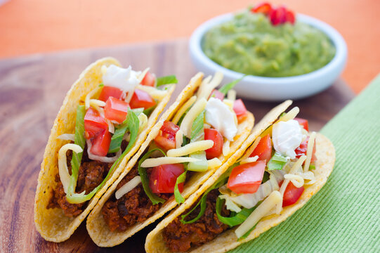 Tacos with beef in hard shells with lettuce, tomato, cheese and guacamole in background, close up