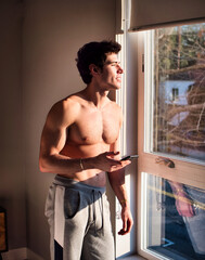 Fototapeta na wymiar Young man standing shirtless holding a cell phone, in his bedroom next to window