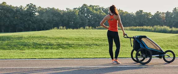 Deurstickers Active mother in sportswear uses jogging stroller for running at park. Happy woman having fun while workout outdoor. Jogging stroller advertisement © Peakstock