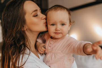 Portrait of woman in white home clothes and her blue-eyed baby. Lady lovingly kisses her daughter