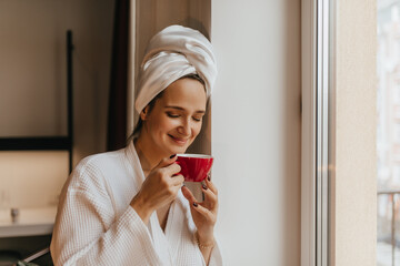 Smiling lady in her morning robe inhales with pleasure aroma of coffee. Woman in white towel on her head holding red cup