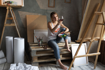 Fototapeta na wymiar beautiful Caucasian artist looking at an abstract painting and holding a glass of wine, sitting in a studio or in a workshop in front of an easel. Young woman doing creativity drinks alcohol.
