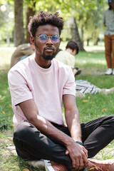 Naklejka na ściany i meble Portrait of African young man in eyeglasses looking at camera while sitting on the grass in the park
