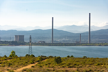 Two refrigeration towers and other chimneys from a power station next to a lake with some mountains on the background
