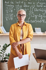 Portrait of confident teacher in eyeglasses standing in the classroom with blackboard in the...