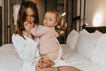 Green-eyed woman in bathrobe kisses hand of her child. Mom and daughter pose at home sitting on bed