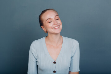 Portrait of young smiling european woman closed eyes. Beautiful happy blonde girl wears t-shirt and looks at camera. Studio shoot isolated on blue background.