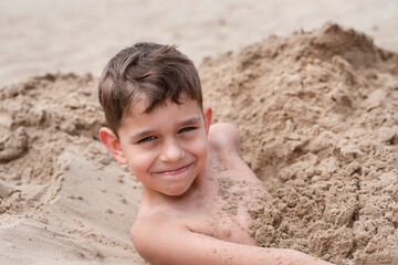 A young boy resting on the beach almost completely buried in the sand and sincerely smiling, happy...
